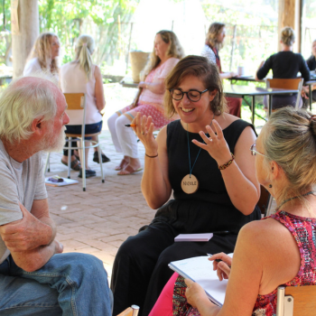 Three people seated in conversation at an outdoor gathering, with others talking at tables in the background.